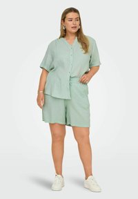 Světle zelený dvoudílný outfit obsahující košili s krátkým rukávem a knoflíky a odpovídající šortky s elastickým pasem. Měkká textilie.