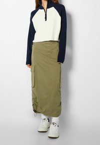 Skuren, krämig och marinblå ribbad tröja med hög krage; lång, olivgrön cargokjol med fickor; vita sneakers med bruna detaljer.