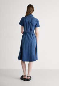Denim shirt dress in a medium blue wash with short sleeves and a belted waist. Features a flared skirt and a collar with stitching details.