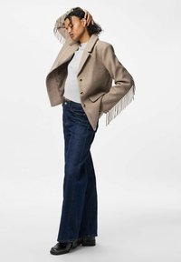 Franjeeritud, beež villajakk, millel on nurgeline kaelus, ühe nööbiga kinnitamine, millele on kombineeritud tume sinine laiadest jalad denimiste teksad ja mustad pahkluudel viikingisaapad.