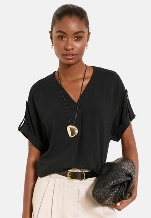 Žena v čiernej blúzke s výstrihom do V, béžové plisované nohavice, čierny opasok, zlatý prívesok na náhrdelníku, drží čiernu tkanú kabelku na zápästie.