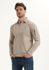 Beżowa koszulka polo z długim rękawem, z kołnierzykiem, trzema guzikami i małą naszywką z logo. Materiał ma gładką teksturę, w zestawieniu z niebieskimi dżinsami.