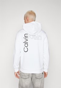 Bílá mikina s kapucí má na zadní straně vertikální černé logo "Calvin Klein". Měkký materiál s klasickým střihem a žebrovanými manžetami.