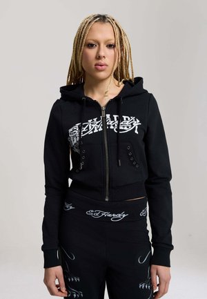 Ung person med fletninger iført en sort Ed Hardy lynlåshoodie og matchende bukser med hvidt logo og grafiske designs.