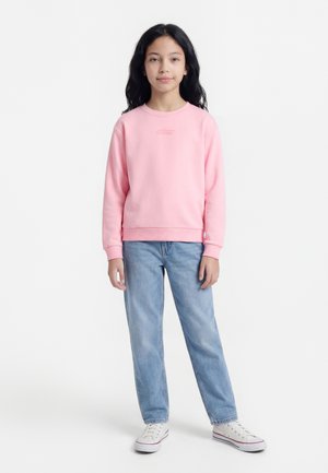 Mladé dievča s dlhými čiernymi vlasmi, oblečené v ružovej mikine, svetlomodrých džínsoch a bielych teniskách, stojí pred bielym pozadím.