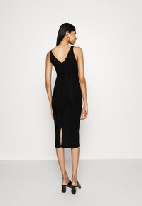 Black knitted midi dress with a V-back, ribbed texture, side slit, and fine detailing. Worn with open-toe heeled shoes.
