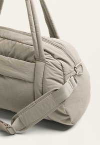 Vaalea beige pehmeä matkakassi, jossa on kaksi tukevuutta käsinojaa, säädettävä olkahihna ja vetoketjukiinnitys. Teksturoidusta kankaasta valmistettu, jossa näkyvät ompelutiedot.