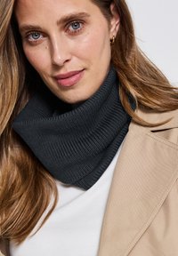 Collo grigio scuro lavorato a maglia, con una texture a coste. Indossato con un cappotto beige sopra una maglietta bianca, evidenziando versatilità e calore.