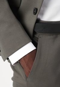 Costume gris en tissu texturé, ceinture en cuir noir et une manche de chemise blanche qui dépasse. La main, de couleur marron, repose dans la poche.