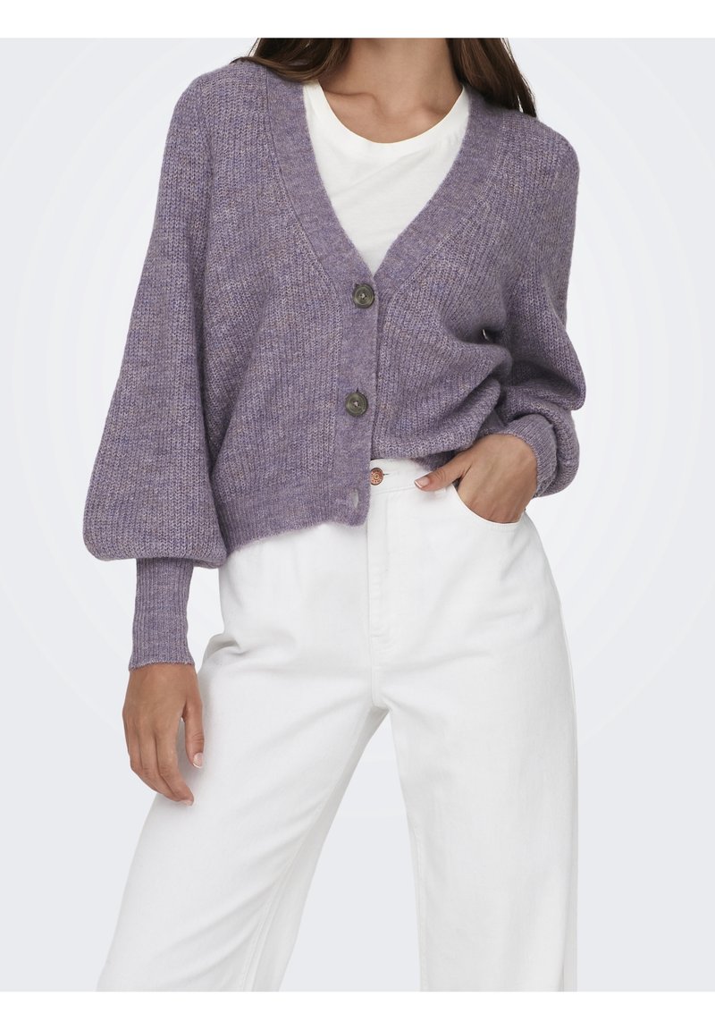 Femme portant un cardigan lavande à col en V boutonné avec des manches ballon par-dessus une chemise blanche, associé à un pantalon blanc taille haute.