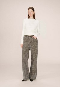 Femme debout, portant un haut à manches longues blanc, un jean large à taille haute avec un motif léopard, et des chaussures à talons noires.