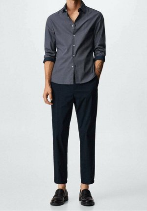 Man draagt slanke marineblauwe broek, zwarte leren loafers en een aangeknoopte marineblauwe blouse met kleine witte stippen, mouwen opgerold.