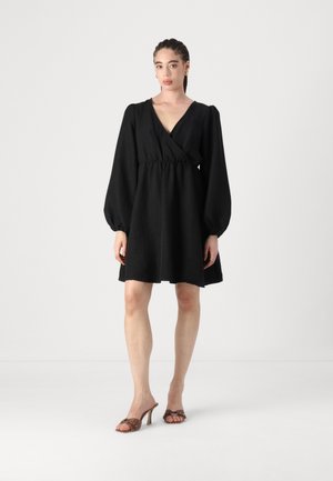 Black wrap dress with voluminous long sleeves and a cinched waist. Smooth texture, knee-length, paired with brown heeled sandals.