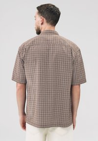 Camisa marrón de manga corta con un patrón de cuadros. Presenta un cuello estándar y un dobladillo recto. La tela parece ligera y texturizada.