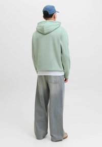 Mintgroene hoodie met een trekkoordcapuchon, gecombineerd met lichtgekleurde, wijd uitlopende jeans. De pet is blauw en de schoenen zijn beige.