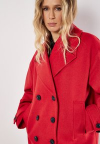 Punainen villa-sekoitteinen kaksirivinen takki, jossa on mustat napit, leveät kaulukselliset läppäkaulukset ja sivutaskut. Pehmeä rakenne ja ylisuurta istuvuutta.