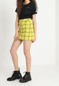 Maglietta nera a maniche corte e una mini gonna a quadri gialla con un design aderente, abbinata a stivaletti neri con lacci alla caviglia.