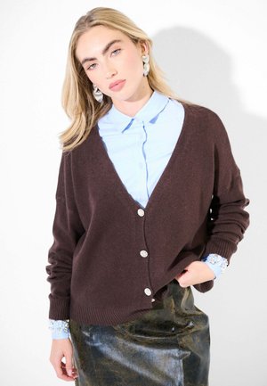 Cardigan marrone lavorato a maglia con scollo a V e chiusura con bottoni lucidi, abbinato a una camicia colletto blu chiaro e una gonna scura testurizzata.
