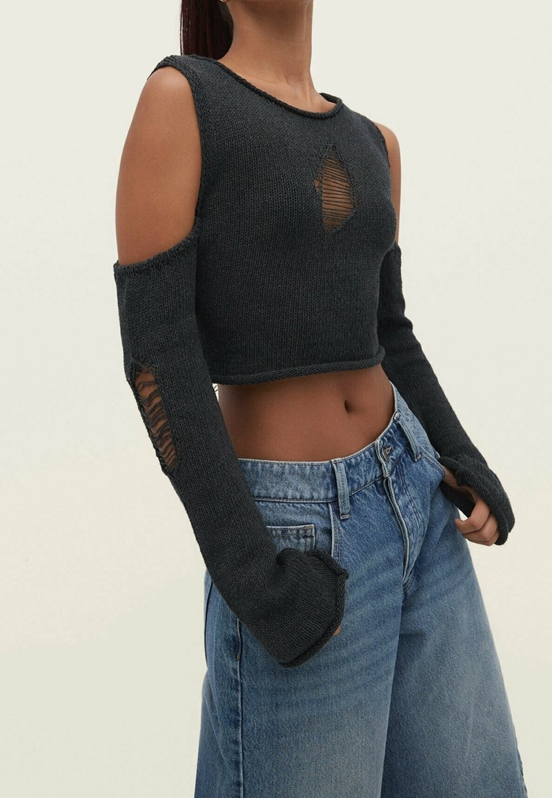 Woman wearing black knit crop top with open shoulders and lace holes on chest and sleeves, paired with high-waisted blue jeans.