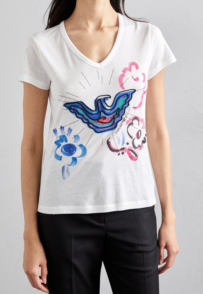Femme portant un t-shirt blanc à col en V avec des motifs abstraits colorés d'oiseaux bleus et roses et de fleurs, assorti à un pantalon noir.