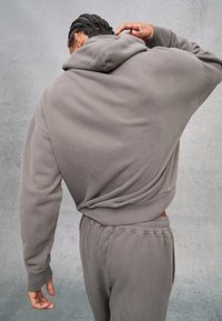 Felpa grigia con cappuccio e pantaloni abbinati in tessuto morbido. La felpa presenta un grande cappuccio e una vestibilità comoda, enfatizzando comfort e semplicità.