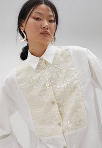 White shirt with a textured cream panel featuring sequins, a pointed collar, and light-colored buttons along the front.