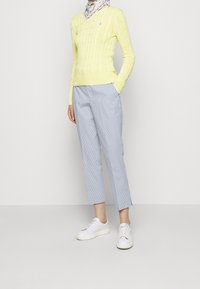 Žlutý svetr s copánkovým vzorem, světle modré pruhované kalhoty a bílé tenisky. Oblečení má texturovaný vzor pletení a ležérní střih.