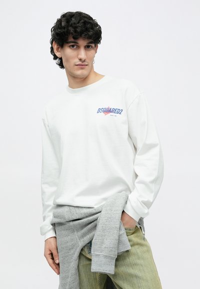 Jauns vīrietis ar cirtainiem matiem, valkājot baltu DSQUARED2 sporta kreklu, gaiši zaļas džinsas un pelēku džemperi, kas sasiets ap vidukli.