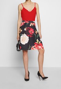 Top a camicia rosso con bordi scallop, vita attillata, gonna nera con grande motivo floreale in rosso e bianco, abbinata a tacchi neri e una piccola borsa.