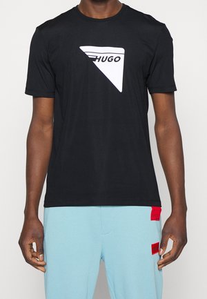 Man draagt een zwart t-shirt met een wit driehoekig "HUGO" logo en lichtblauwe broek met twee rode rechthoekige patches.