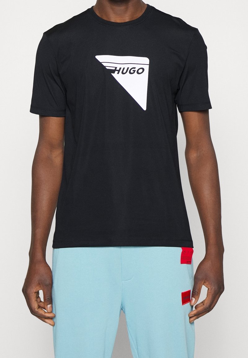 Homme portant un t-shirt noir avec un logo triangulaire blanc "HUGO" et un pantalon bleu clair avec deux patchs rectangulaires rouges.