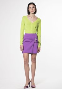 Cardigan verde brillante con scollo a V e bottoni, abbinato a una gonna avvolgente viola con un accento decorativo. Il modello indossa tacchi alti viola.