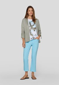 Hellgraue offene Strickjacke, Grafik-T-Shirt in Weiß und Blau und hellblaue, verkürzte Hose. Braune flache Sandalen. Sanfte Texturen, lässige Passform.