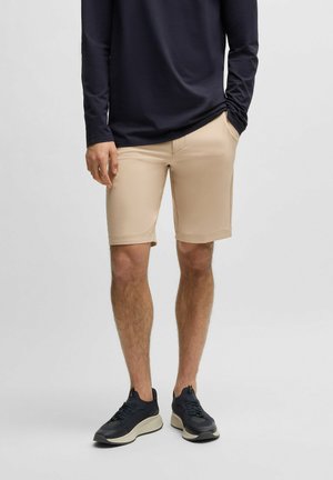 Pantaloni corti color sabbia in tessuto leggero e liscio, con taglio dritto e orli risvoltati, abbinati a una maglietta a maniche lunghe di colore blu scuro e sneakers nere.