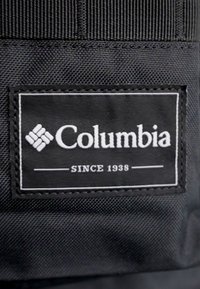 Czarna metka z tkaniny z logotypem "Columbia" w białym kolorze, z napisem "SINCE 1938", z przeszytymi krawędziami i gładką powierzchnią.