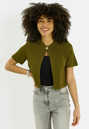 Mujer con cabello rizado sonriendo, con un cárdigan de manga corta color verde oliva sobre una blusa negra, jeans de tiro alto color gris claro y una pulsera plateada.