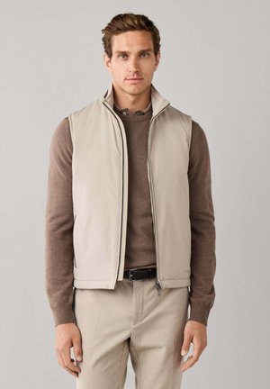Man draagt een beige mouwloos ritjesvest over een bruine trui met lange mouwen en beige broek tegen een effen grijze achtergrond.
