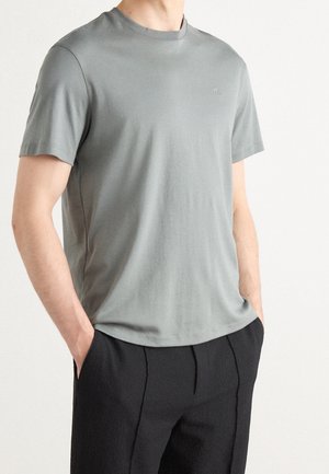 Homme portant un t-shirt gris clair à manches courtes et col rond, légèrement rentré dans un pantalon noir ample et texturé, les mains dans les poches.
