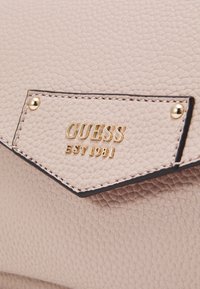 Beige struktureret lædertaske med syet klap, sikret med guldskruer og guld "GUESS EST 1981" præget logo.