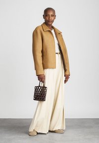 Camel ribbet jakke, hvid top, cremefarvede vide bukser og beige vævede sko. Holder en brun nettaske. Neutral baggrund.