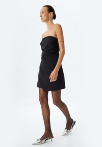 Svart strapless miniklänning med en figurnära design; modellen bär genomskinliga svarta strumpbyxor och silverfärgade stilettklackar med dekorativa detaljer.