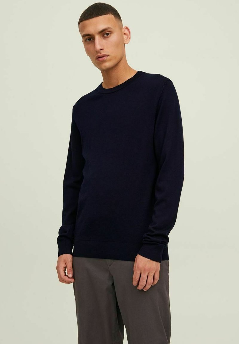 Maglione lavorato a maglia blu navy con scollatura rotonda, maniche lunghe e polsini a coste. Texture morbida e vestibilità classica, abbinato a pantaloni grigi chiari.
