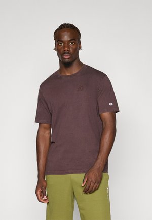 Braunes Baumwoll-T-Shirt mit Rundhalsausschnitt. Verfügt über ein subtils Logo auf der Brust und kurze Ärmel. Kombiniert mit grünen Jogginghosen.