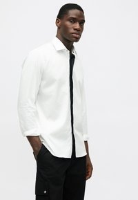 Camisa de manga longa branca com uma pala de botões pretos no centro, punhos enrolados e um corte slim, combinada com calças cargo pretas.