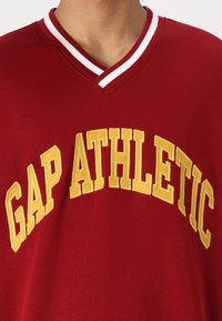 Camiseta de algodón roja con escote en V, que presenta un gran texto bordado en amarillo "GAP ATHLETIC". Detalle de raya blanca en el cuello.