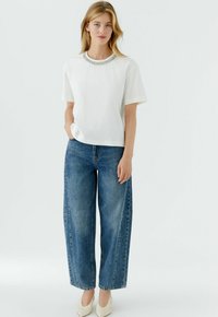 Weißes kurzärmeliges T-Shirt mit verzierendem Ausschnitt, kombiniert mit locker sitzenden blauen Denim-Jeans und cremefarbenen spitzen Schuhen.