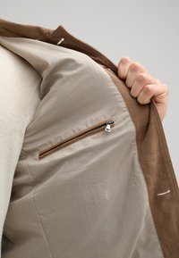 Braune Wildlederjacke mit einem beigen Innenfutter. Sie verfügt über eine Reißverschlusstasche und eine glatte Textur, die die kontrastierenden Stofffarben hervorhebt.