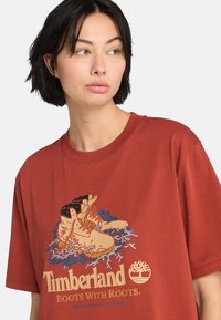 Rød bomulds T-shirt med en grafisk illustration af støvler med rødder, blå bølger og teksten "Timberland Støvler Med Rødder." Korte ærmer.