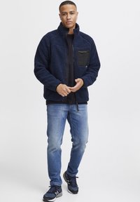 Navy fleece jacket with a black zippered chest pocket, high collar, and soft texture, paired with blue jeans and athletic shoes.
