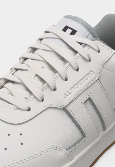 Λευκό δερμάτινο αθλητικό παπούτσι με λευκά κορδόνια, ραμμένες λεπτομέρειες, οπές κοντά στα δάχτυλα και μερικώς ορατή γκρι εσωτερική επένδυση.